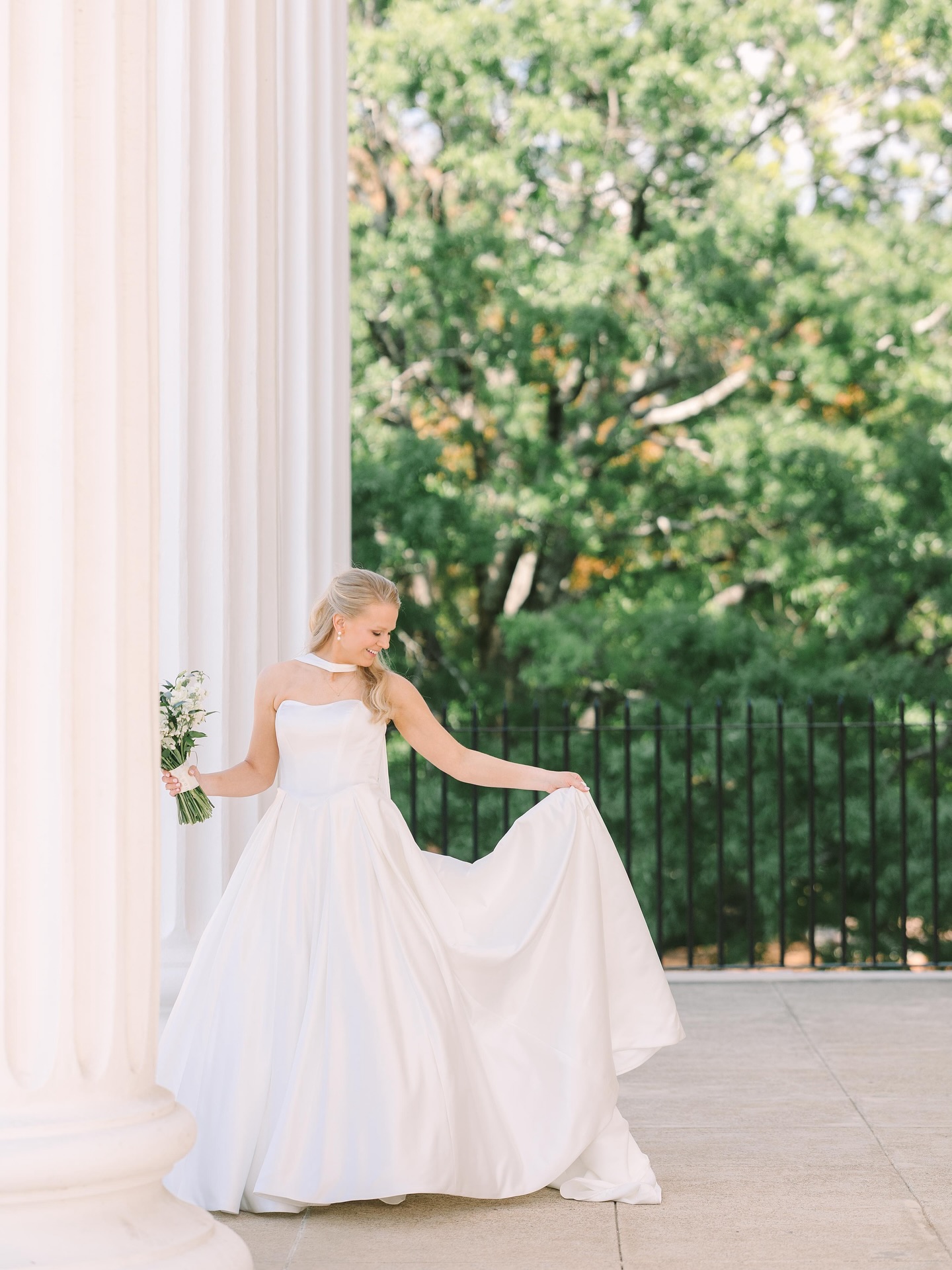 Columbia SC Wedding Photographers | Stee Garman Photography Fine-art Wedding Photographer | A bride in a flowing white gown stands gracefully by white columns, holding a bouquet. Sunlight filters through trees, creating a serene, elegant atmosphere.