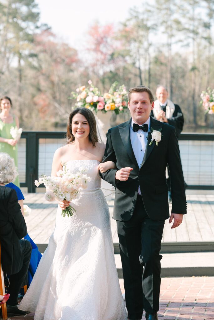 Outdoor Wedding Venues Columbia SC | Stee Garman Photography Fine-art Wedding Photographer | A couple in formal attire walks down an outdoor aisle. The bride holds a bouquet, wearing a white gown, while the groom is in a black tuxedo. Smiling guests and floral arrangements are visible in the background.