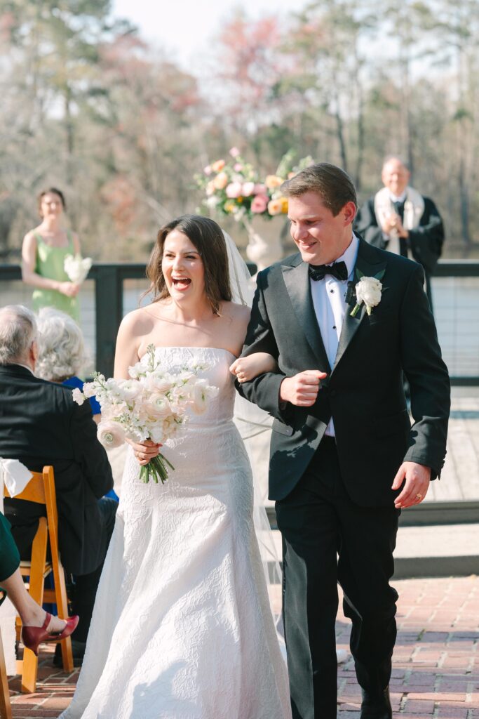 Wedding Photographer Columbia SC | Stee Garman Photography Fine-art Wedding Photographer | Bride in white dress and groom in tuxedo joyfully walk down the aisle, arm in arm, outdoors. Guests seated, flowers and greenery in background.
