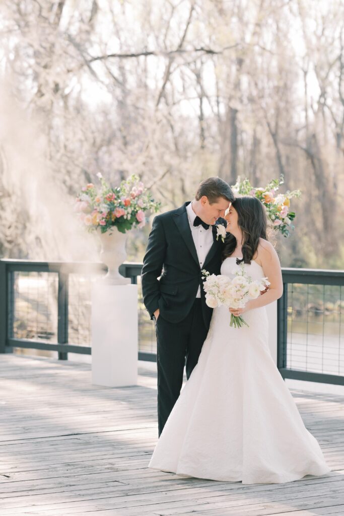 Wedding Planning in Columbia SC | Stee Garman Photography Fine-art Wedding Photographer | A bride in a white gown and a groom in a black suit stand on a wooden deck, looking at each other. She holds a bouquet of white flowers. Trees with spring blossoms are in the background.