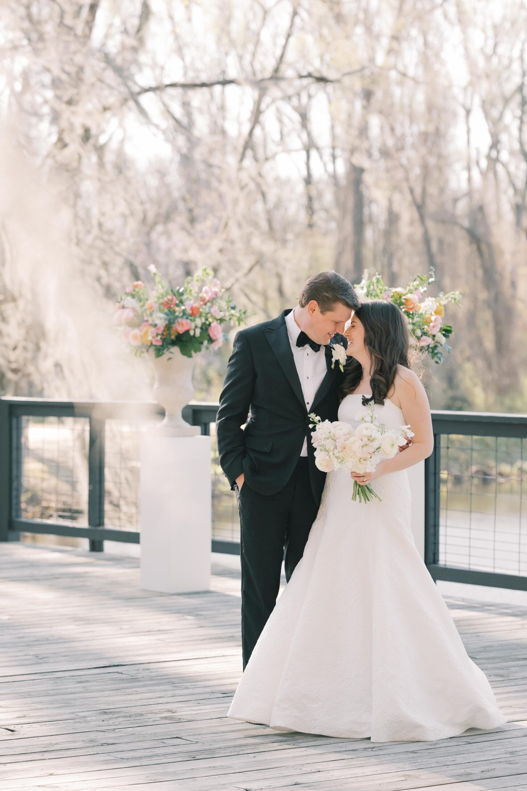 Wedding Photographer Columbia SC | Stee Garman Photography Fine-art Wedding Photographer | A bride in a white gown and a groom in a black suit stand on a wooden deck, looking at each other. She holds a bouquet of white flowers. Trees with spring blossoms are in the background.
