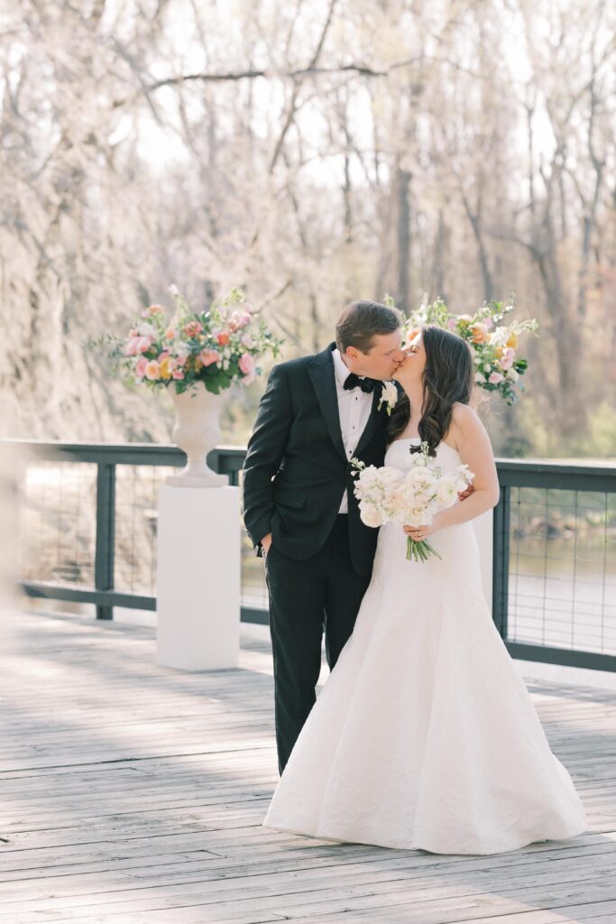 Wedding Photographer Columbia SC | Stee Garman Photography Fine-art Wedding Photographer | A bride in a white gown and a groom in a black suit stand on a wooden deck, kissing. She holds a bouquet of white flowers. Trees with spring blossoms are in the background.