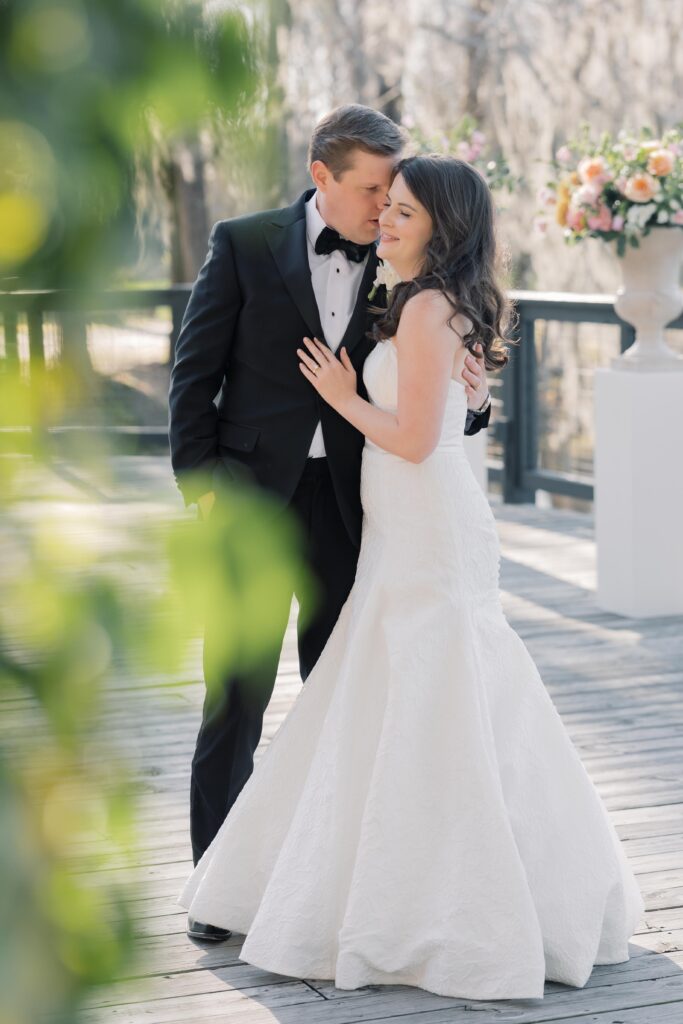 Wedding Photographer Columbia SC | Stee Garman Photography Fine-art Wedding Photographer | Bride and groom embrace on a sunlit wooden deck. Bride in a flowing white gown, groom in a black tuxedo. Soft-focus flowers and greenery create a romantic tone.
