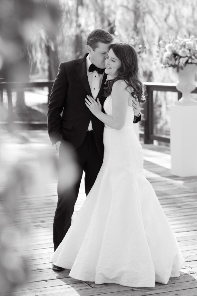 Columbia SC Wedding Photographers | Stee Garman Photography Fine-art Wedding Photographer | A bride in a white gown and a groom in a tuxedo embrace lovingly on a wooden deck, with soft-focus flowers and trees in the background, conveying romance.