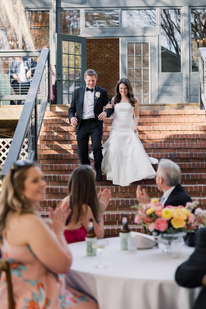 Wedding Photographer Columbia SC | Stee Garman Photography Fine-art Wedding Photographer | Bride in a white gown and groom in a tuxedo descend brick stairs at a wedding, guests clapping and smiling at a flower-adorned table below.