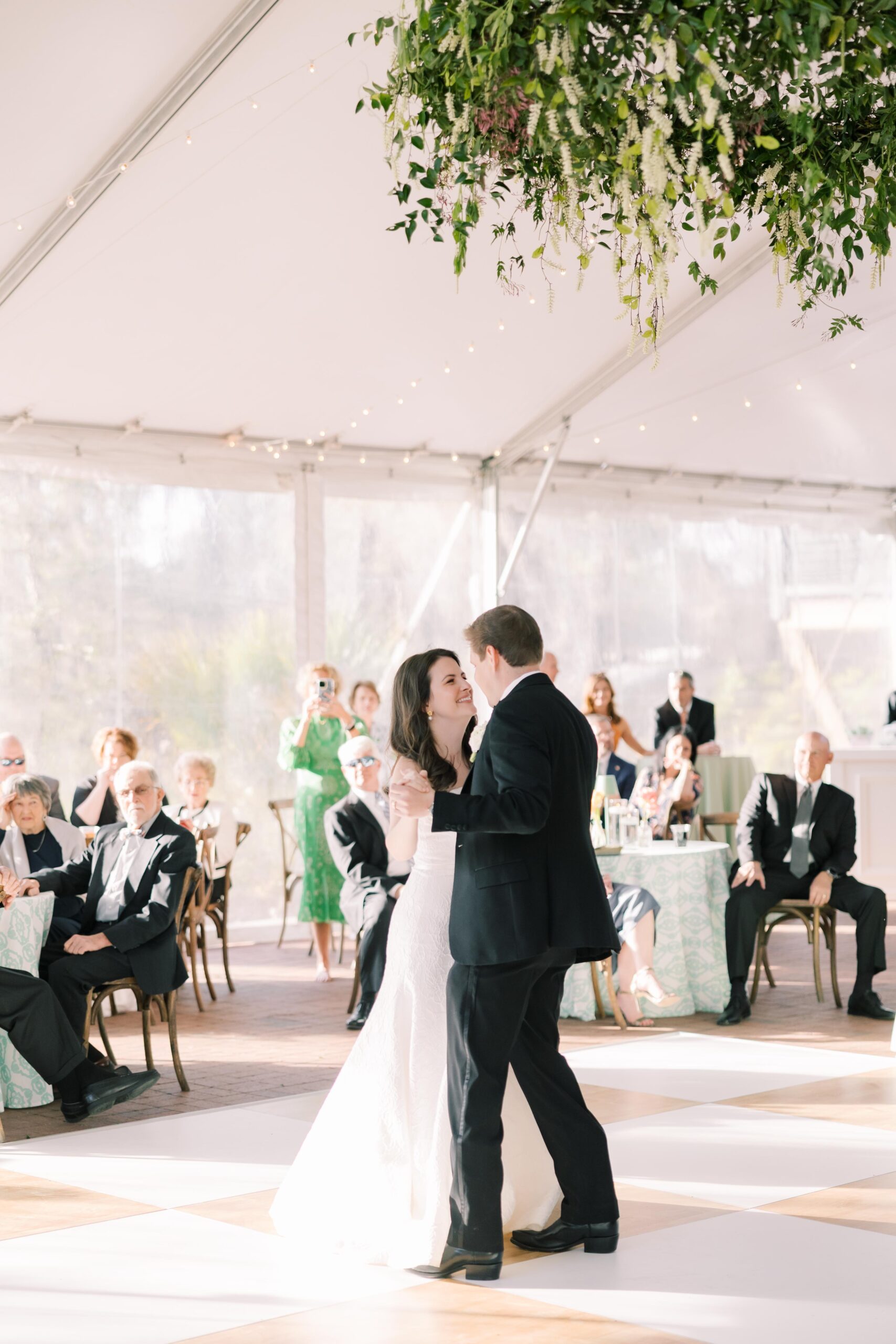 Wedding Reception Venues in Columbia SC | Stee Garman Photography Fine-art Wedding Photographer | A couple shares a joyful first dance at a wedding reception under a tent adorned with greenery. Guests in formal attire watch and smile warmly.