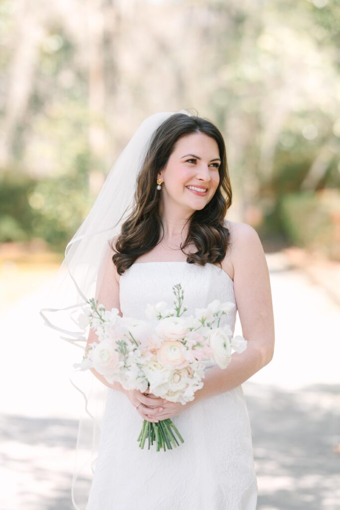 Wedding Photographer Columbia SC | Stee Garman Photography Fine-art Wedding Photographer | Bride in a strapless white gown and veil smiles while holding a bouquet of pale flowers. She stands on a sunlit path with blurred greenery behind.