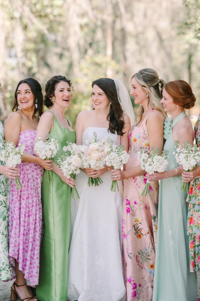 Wedding Photographer Columbia SC | Stee Garman Photography Fine-art Wedding Photographer | A bride in a white gown stands with bridesmaids wearing floral and pastel dresses, all holding bouquets. They all smile joyfully.