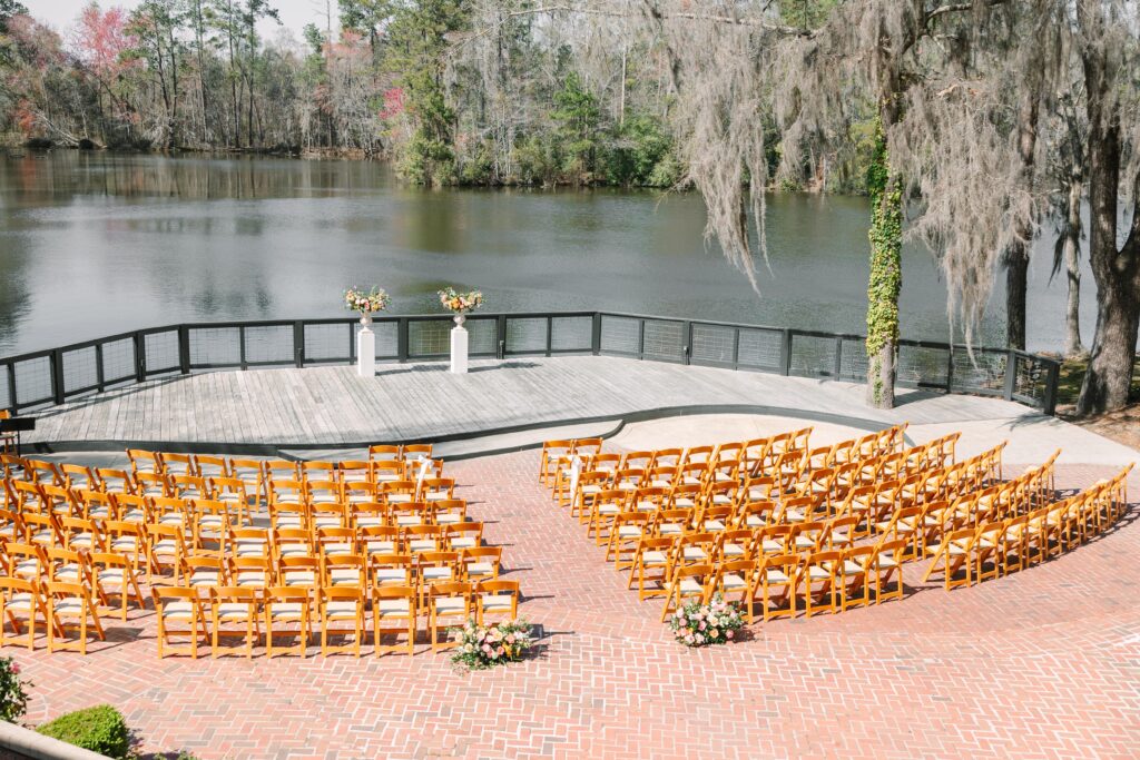 South Carolina Wedding Photographers | Stee Garman Photography Fine-art Wedding Photographer | Sunlit outdoor wedding setup with rows of wooden chairs on a brick patio facing a lakeside platform adorned with floral arrangements, surrounded by trees.
