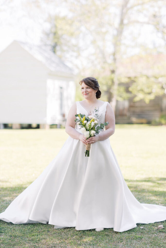 Wedding Venues in Columbia SC | Stee Garman Photography Fine-art Wedding Photographer | Bride in a flowing white gown stands outdoors, holding a bouquet of yellow and white flowers. She smiles softly with a serene, sunny backdrop.