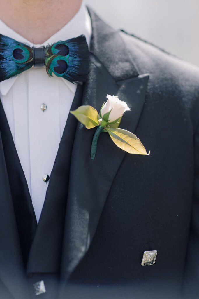 Wedding Venues in Columbia SC | Stee Garman Photography Fine-art Wedding Photographer | Close-up of a man in a black tuxedo wearing a vibrant peacock feather bow tie. A white rose boutonniere is pinned to his lapel. Formal and elegant tone.
