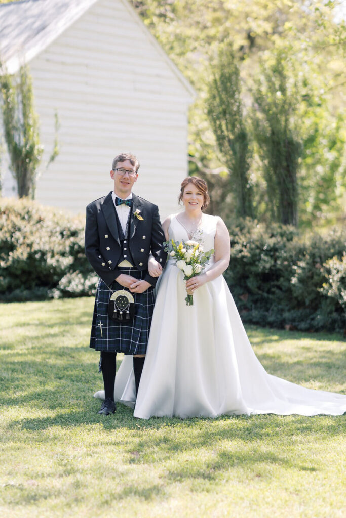 Wedding Venues in Columbia SC | Stee Garman Photography Fine-art Wedding Photographer | A bride and groom stand on grass in front of a white building. The groom wears a kilt and tuxedo jacket, while the bride wears a white gown, holding yellow flowers.