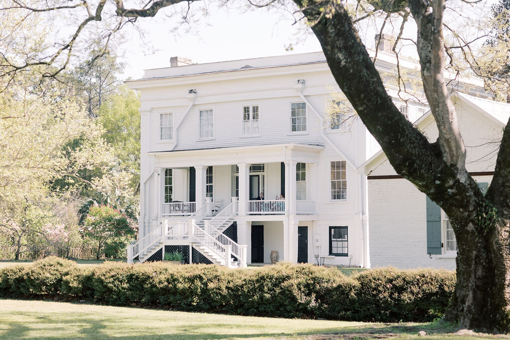 The white Wavering Place wedding venue sits beneath tall trees with manicured hedges, a covered porch, and symmetrical exterior staircases framing the entrance. Its classic Southern setting makes it a distinctive choice for couples planning a wedding in Columbia, SC.