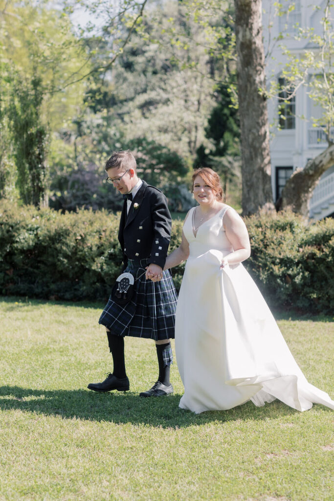 The newlyweds walk hand in hand across the grass as the house peeks through the trees, a graceful portrait scene at Wavering Place Plantation.