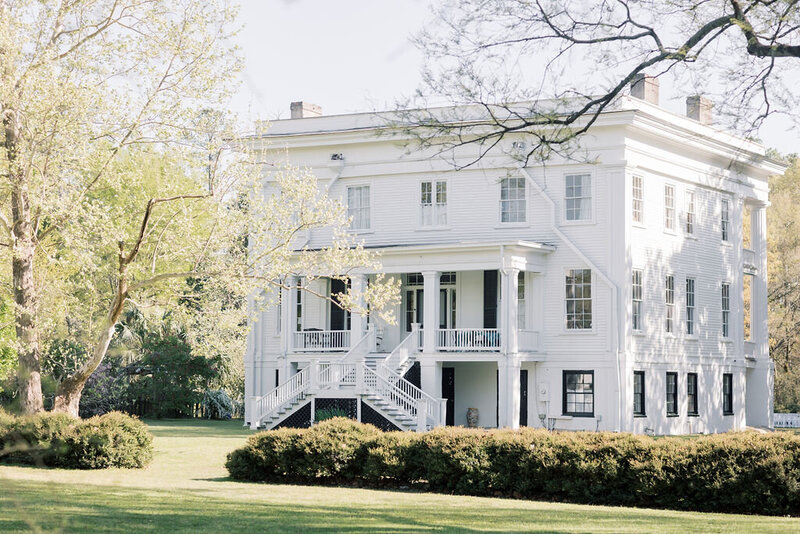 Columbia SC Wedding Photographers | Stee Garman Photography Fine-art Wedding Photographer | A large white colonial-style house with black shutters sits amidst a lush green lawn and tall trees, exuding a peaceful, classic charm.