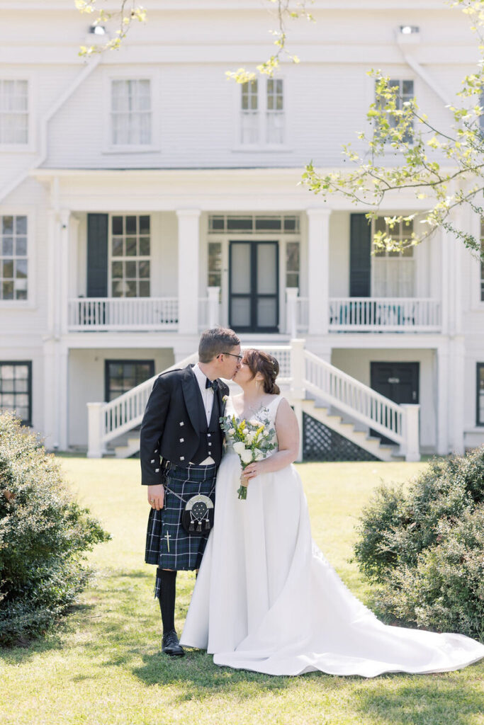 Columbia SC Wedding Photographers | Stee Garman Photography Fine-art Wedding Photographer | A couple shares a kiss in a garden. The groom wears a kilt and the bride a white gown with a bouquet. A white historic house in Wavering Place is in the background.