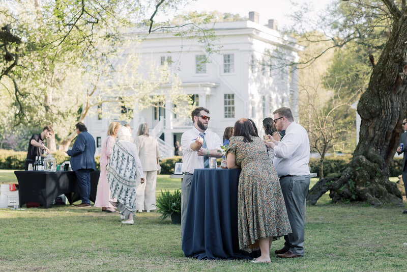 Wedding Reception Venues in Columbia SC | Stee Garman Photography Fine-art Wedding Photographer | People gather around a table with a navy tablecloth at an outdoor event. They are in front of a grand white house, enjoying a sunny day and socializing.