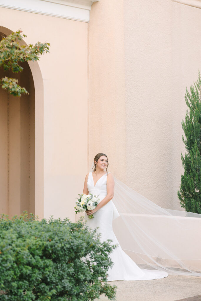 Photography Columbia SC | Stee Garman Photography Fine-art Wedding Photographer | A bride in a white dress and long veil stands smiling, holding a bouquet of white flowers. She's in front of a peach-colored wall with greenery around.