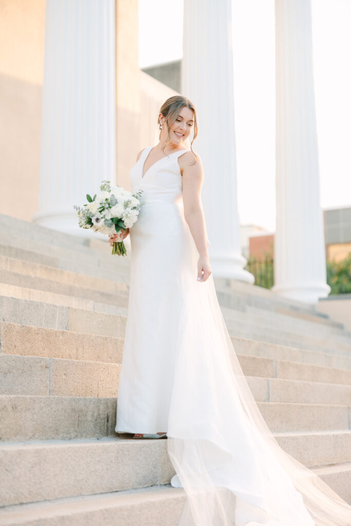 Columbia SC Photography | Stee Garman Photography Fine-art Wedding Photographer | Bride in elegant white gown holding a bouquet stands on stone steps, smiling. Sunlight highlights her and white columns create a classic backdrop.