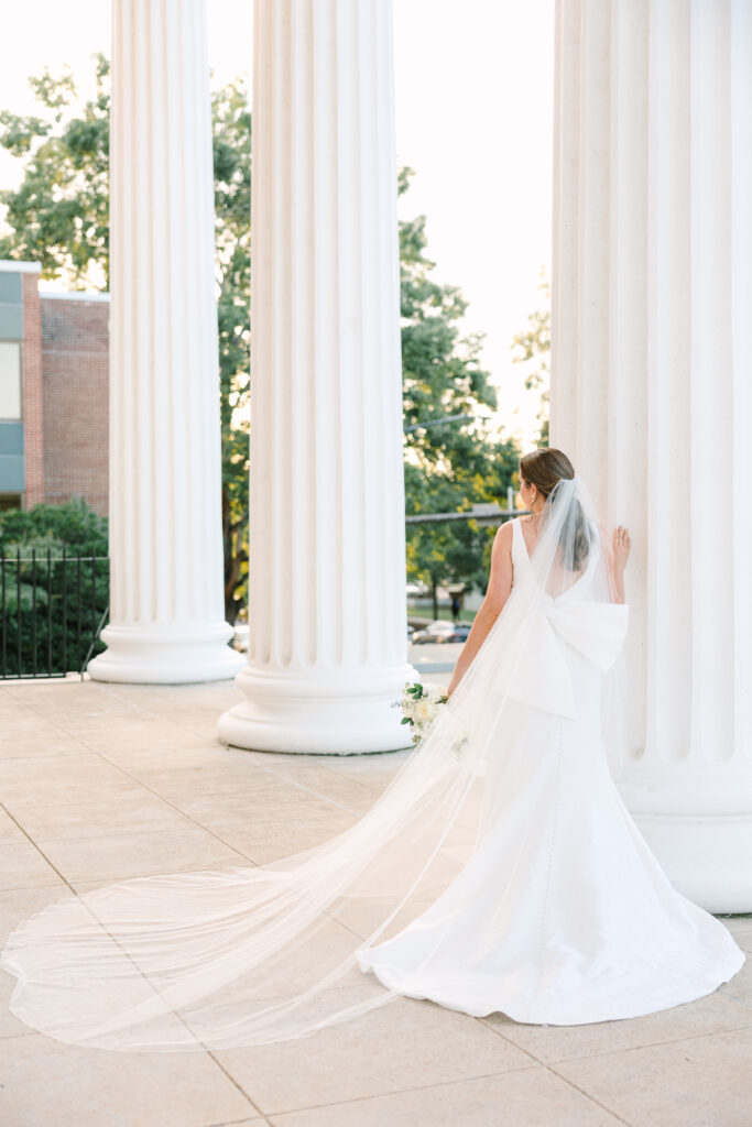 Wedding Venues in Columbia SC | Stee Garman Photography Fine-art Wedding Photographer | Bride in elegant white gown and veil stands by large, white columns. The setting is serene, with soft lighting and lush greenery in the background.