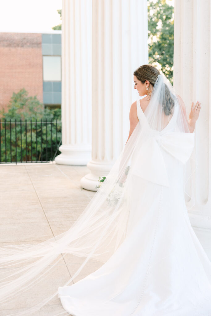 A bride stands between tall white columns with her veil flowing behind her, creating a timeless portrait aligned with classic wedding venues near Columbia SC.
