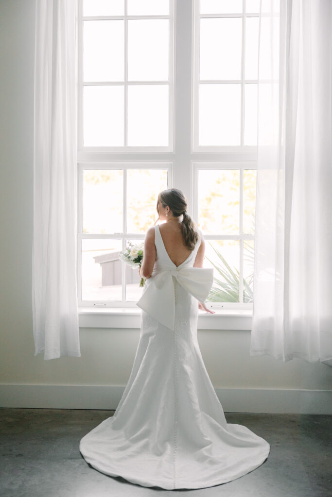 Wedding Venues Columbia SC | Stee Garman Photography Fine-art Wedding Photographer | Bride in a white gown with a large bow stands facing a bright window, holding a bouquet. The soft, natural light creates an elegant, serene atmosphere.