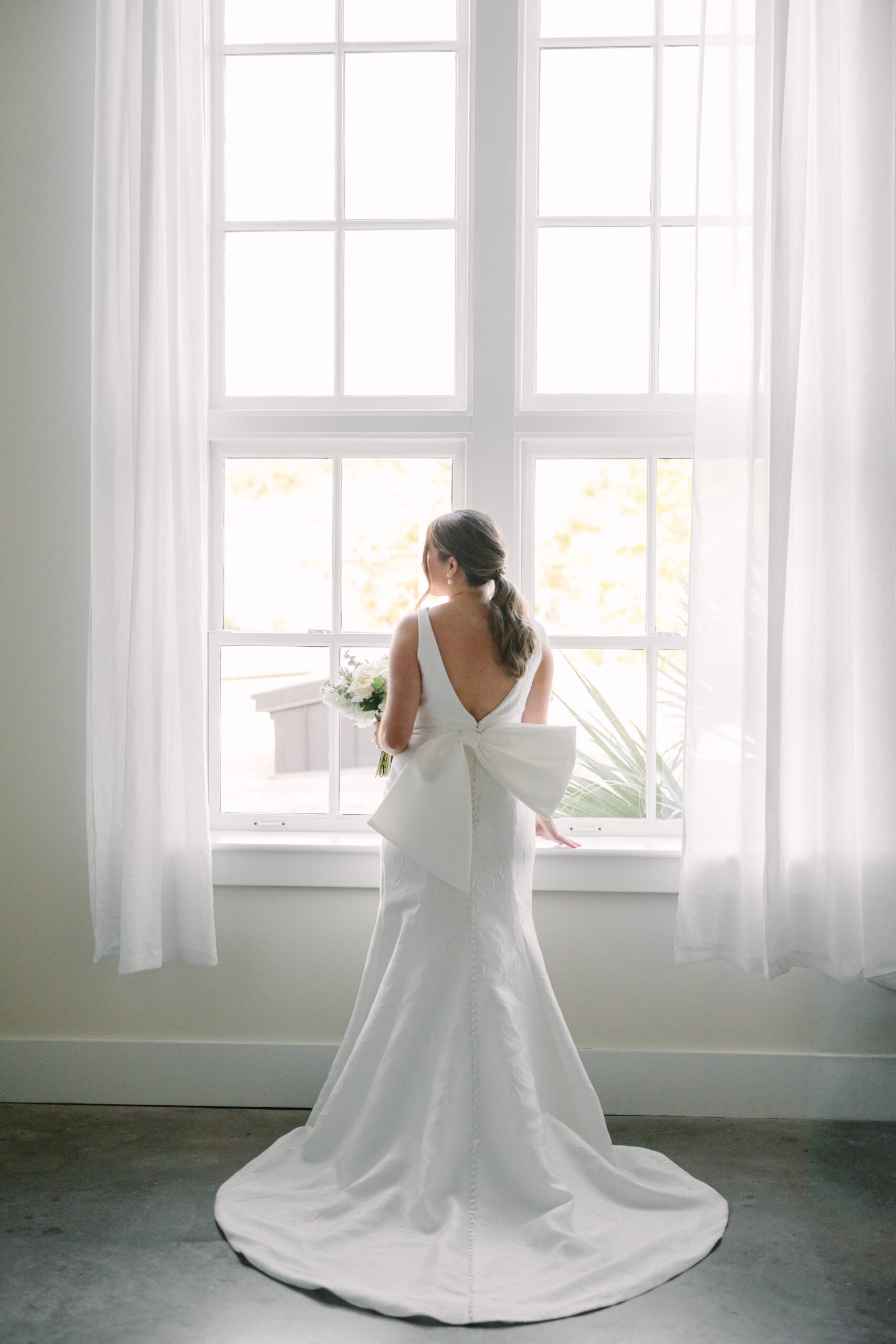 Wedding Venues Columbia SC | Stee Garman Photography Fine-art Wedding Photographer | Bride in a white gown with a large bow stands facing a bright window, holding a bouquet. The soft, natural light creates an elegant, serene atmosphere.