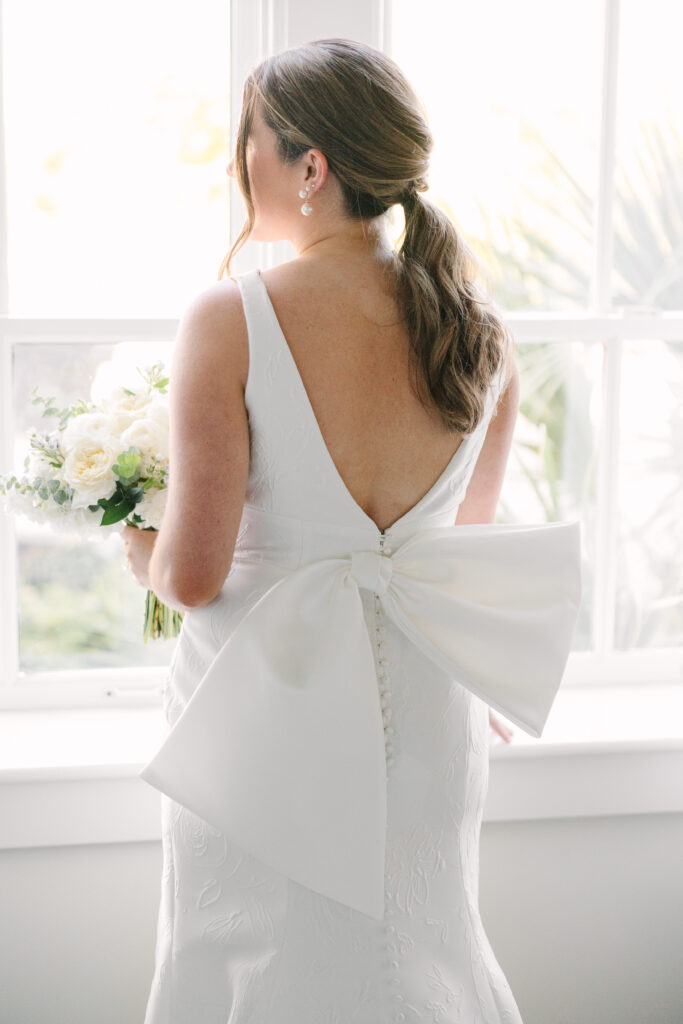 Bridal Portraits Photographer Columbia SC | Stee Garman Photography Fine-art Wedding Photographer | Bride in a white dress with a large bow at the back, holding a bouquet of white roses. She stands by a bright window, creating a serene atmosphere.