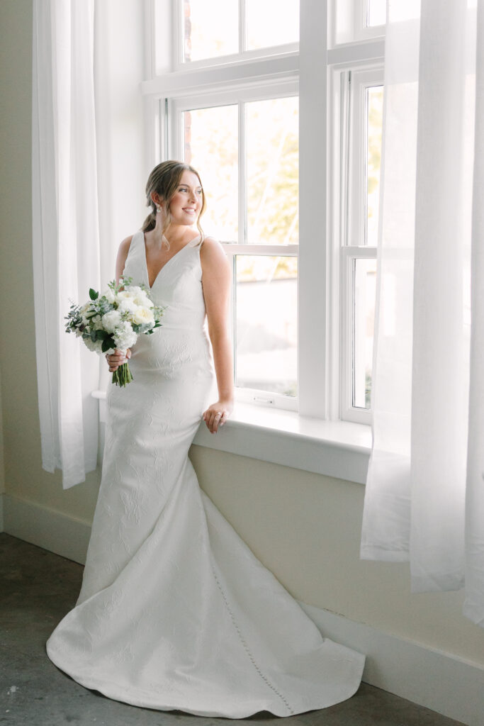 Wedding Venues Columbia SC | Stee Garman Photography Fine-art Wedding Photographer | A bride in a white gown holds a bouquet, leaning against a bright window. Her expression is serene, with soft natural light creating a peaceful ambiance.