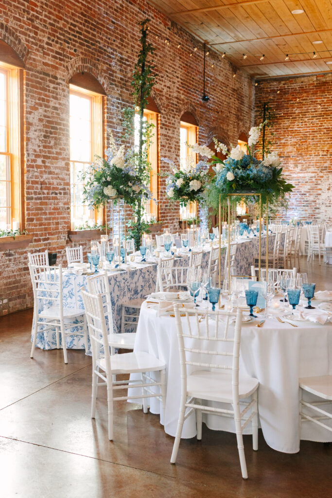 Wedding Reception Venues in Columbia SC | Stee Garman Photography Fine-art Wedding Photographer | Elegant banquet hall with exposed brick walls and tall windows. Tables are set with white linens, blue-patterned cloths, and blue glassware, adorned with lush floral centerpieces.