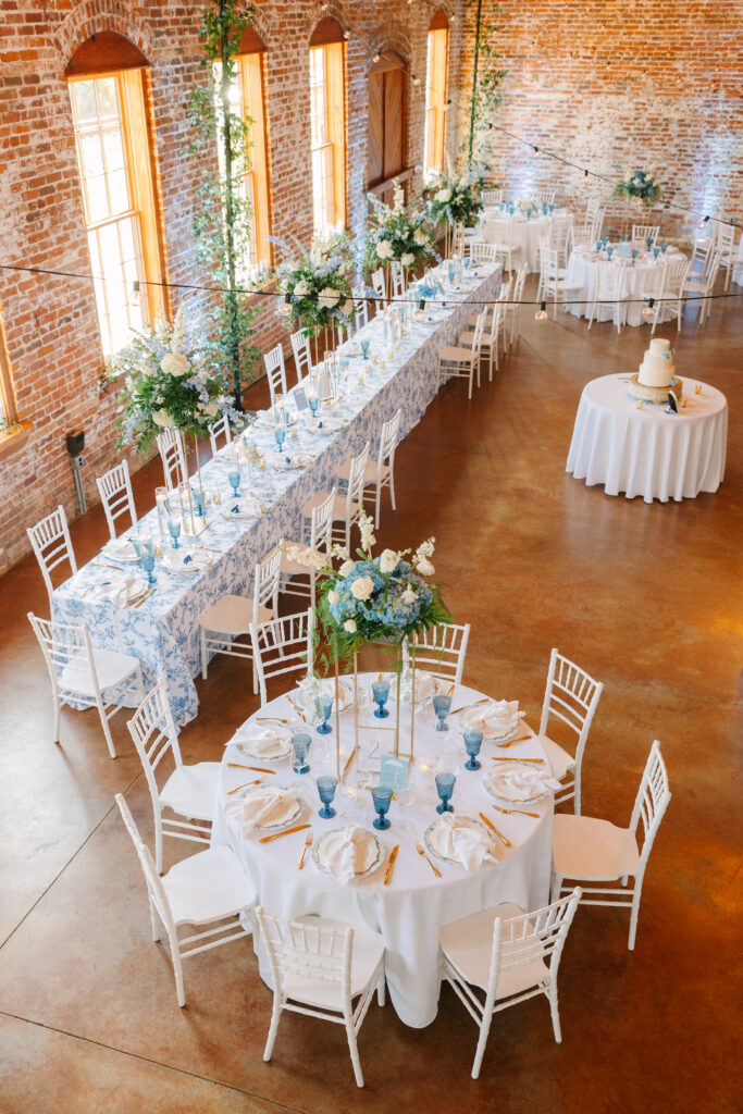 An indoor reception space with exposed brick walls and long tables styled with florals and blue glassware highlights the atmosphere of refined wedding venues in Columbia SC.