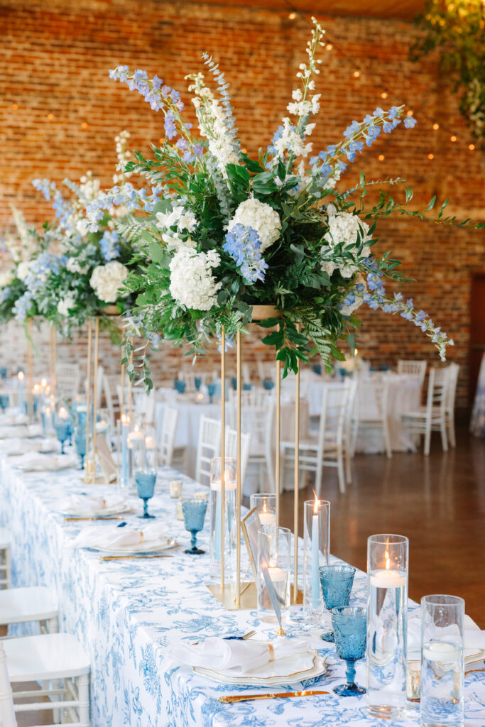 Wedding Venues in Columbia SC | Stee Garman Photography Fine-art Wedding Photographer | Elegant table setting with tall floral centerpieces of white and blue flowers on gold stands. Blue glassware, candles, and a brick wall backdrop add warmth.