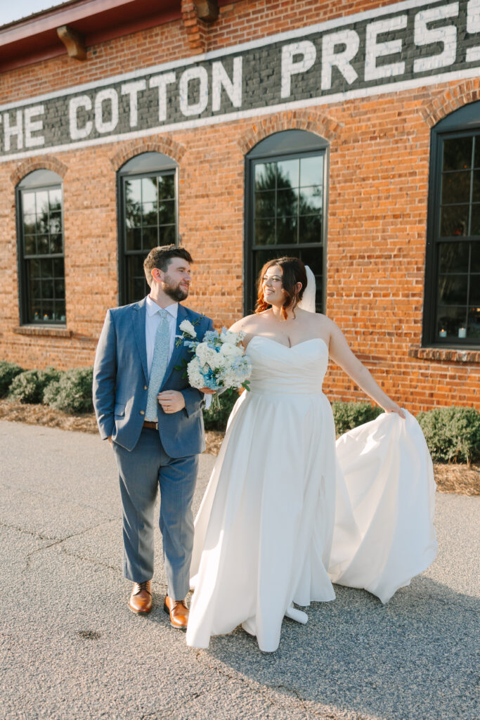 Wedding Venues in Columbia SC | Stee Garman Photography Fine-art Wedding Photographer | A bride and groom happily walk outside a rustic brick building with "The Cotton Press" sign. The bride holds a bouquet and her dress; the groom wears a blue suit.