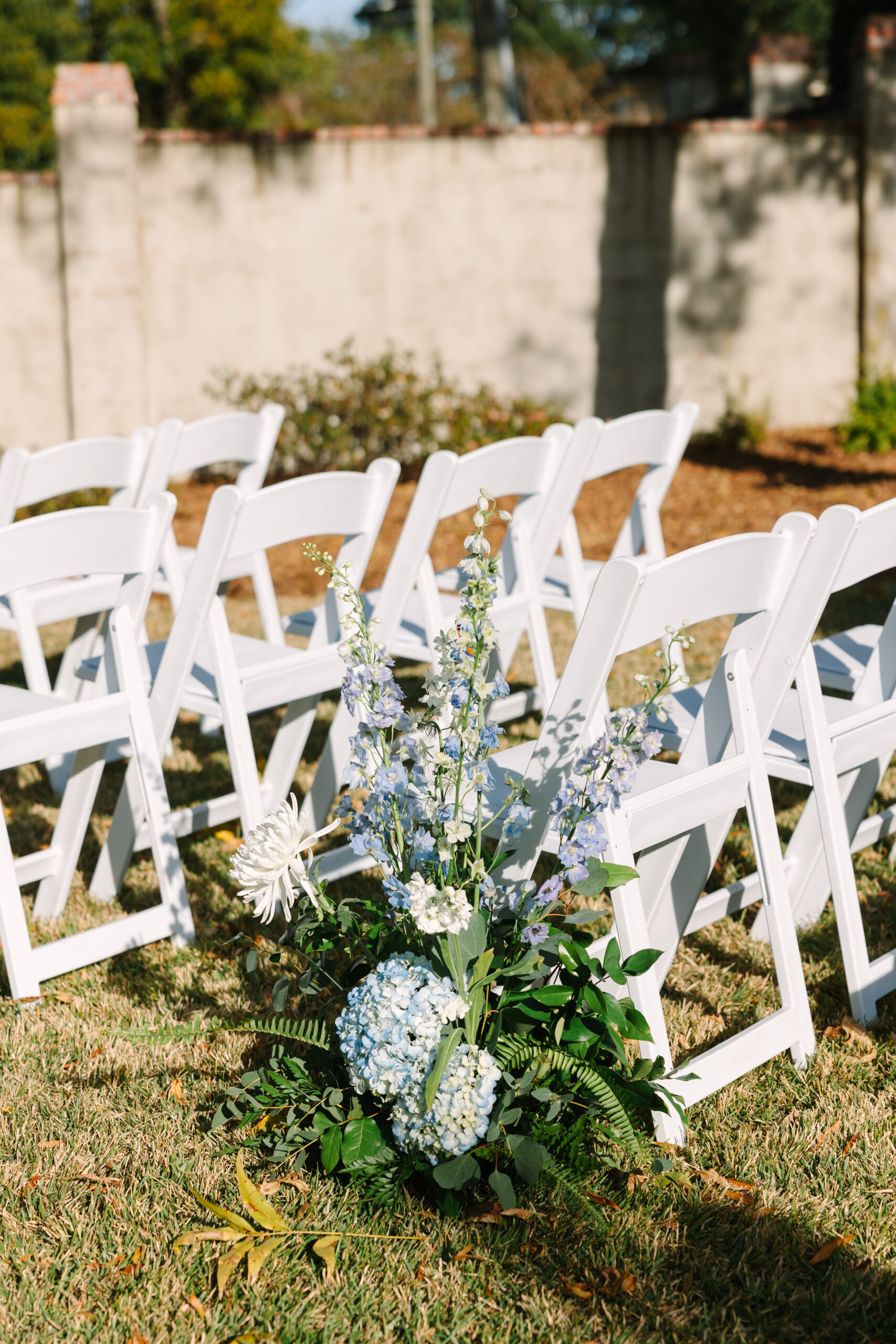 Wedding Venues in Columbia SC | Stee Garman Photography Fine-art Wedding Photographer | Outdoor wedding setting with rows of white folding chairs on grass. A floral arrangement with blue and white flowers and green leaves is in the foreground.