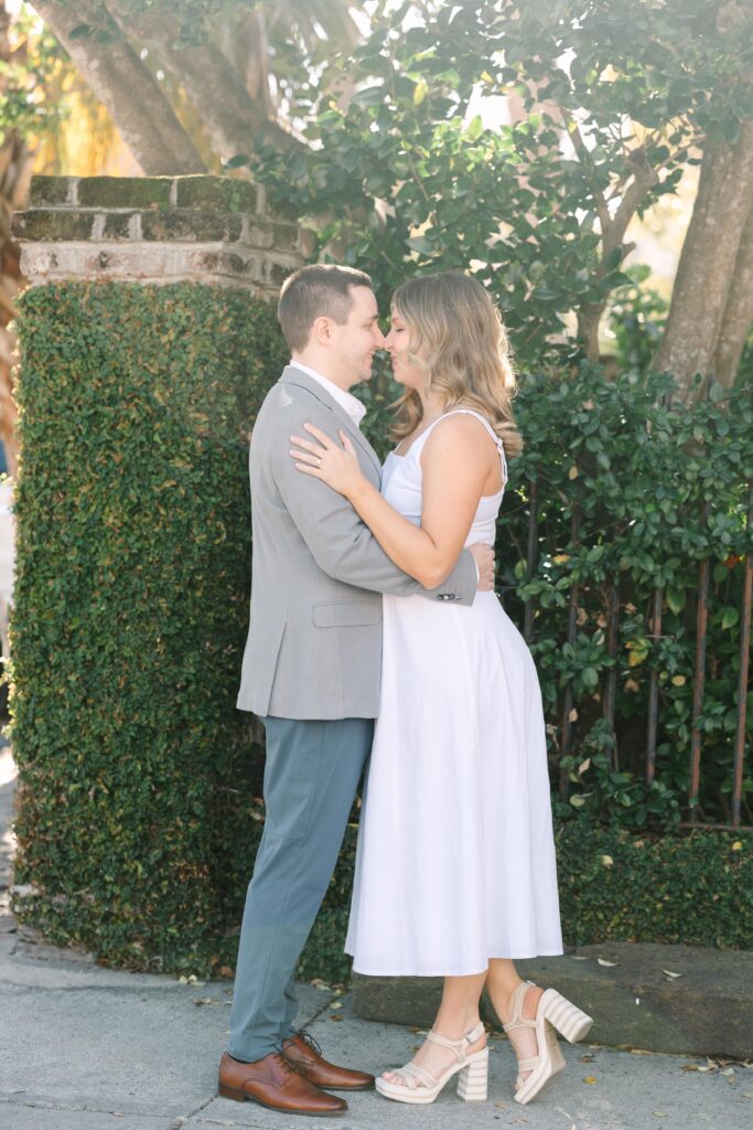 Engagement Photo Locations Columbia SC | Stee Garman Photography Fine-art Wedding Photographer | A couple stands on a white bridge over a calm lake, surrounded by green foliage and vibrant pink flowers. The scene conveys a serene, romantic atmosphere.