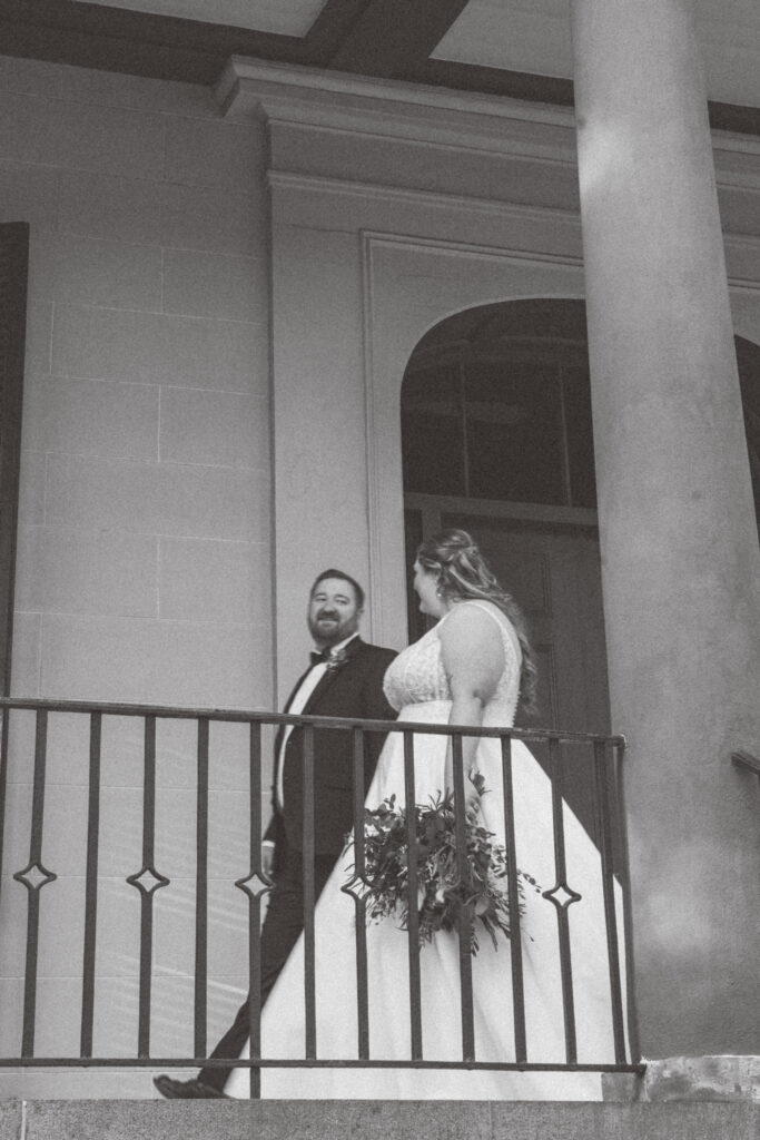 Wedding Venues in Columbia SC | Stee Garman Photography Fine-art Wedding Photographer | A couple stands on a balcony in formal attire, the woman in a white dress holding a bouquet. The man smiles, creating a joyful, romantic atmosphere.