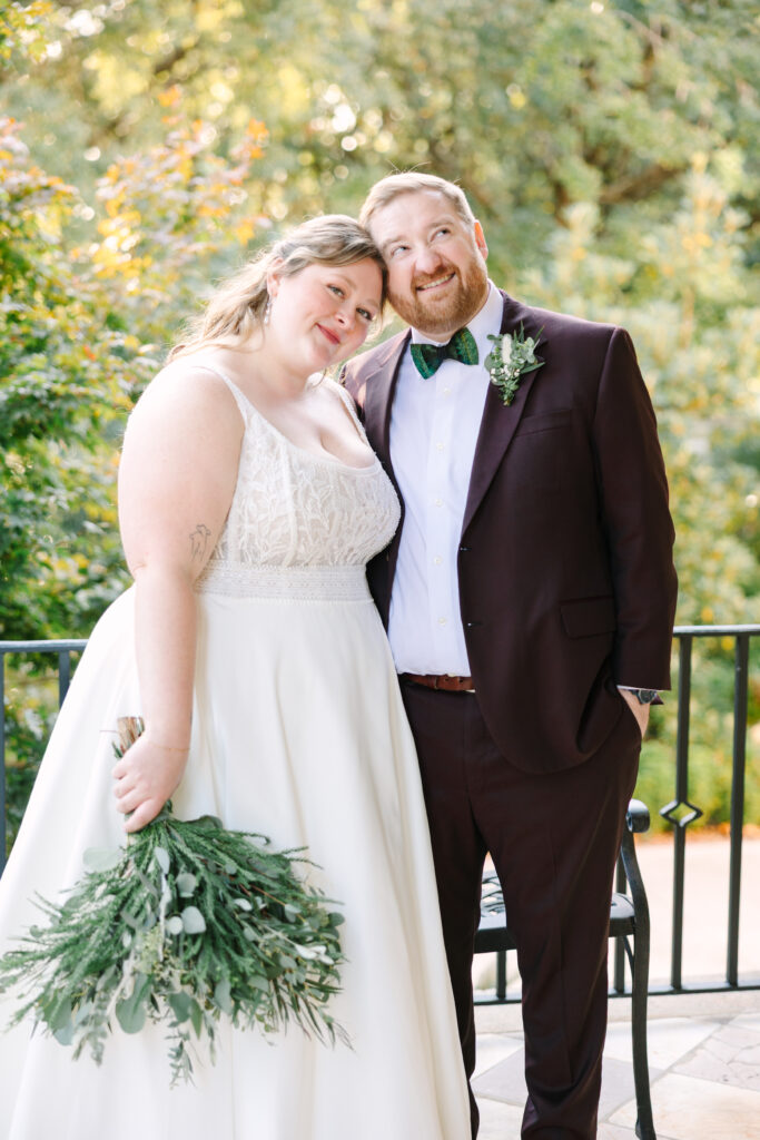 Hampton Preston Mansion Wedding | Stee Garman Photography Fine-art Wedding Photographer | Bride in a white dress and groom in a dark suit with a green bow tie smile lovingly, standing close outdoors against a backdrop of lush greenery.
