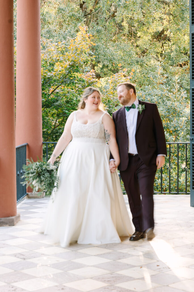 Wedding Venues Columbia SC | Stee Garman Photography Fine-art Wedding Photographer | A joyful bride and groom hold hands, walking on a checkered terrace. She wears a flowing gown, he wears a suit. Greenery surrounds them, creating a serene atmosphere.