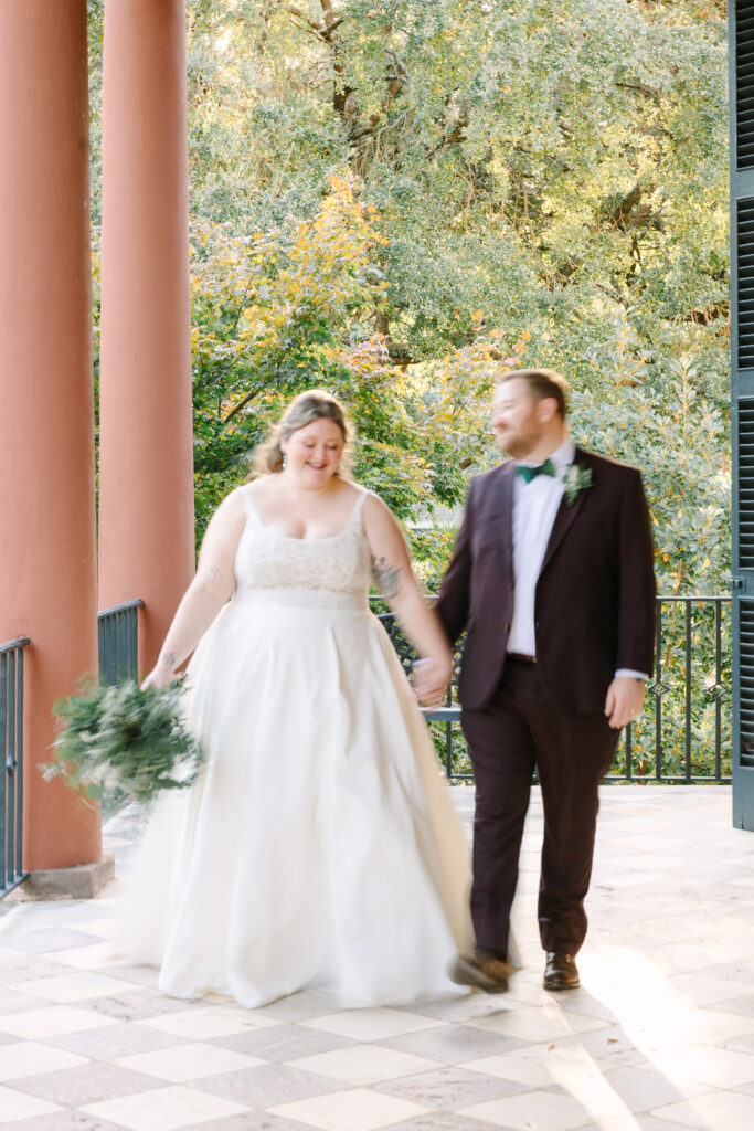 Wedding Planning in Columbia SC | Stee Garman Photography Fine-art Wedding Photographer | A bride in a flowing white gown and a groom in a dark suit walk hand-in-hand on a veranda with columns. They are smiling, surrounded by lush greenery.