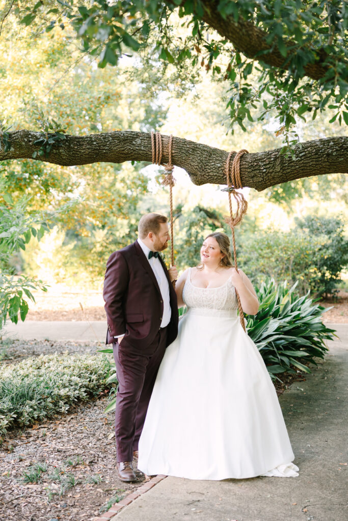 South Carolina Wedding Photographers | Stee Garman Photography Fine-art Wedding Photographer | Bride in a white gown sits on a rustic tree swing, smiling at a groom in a burgundy suit. They stand under a large tree in a sunlit garden.