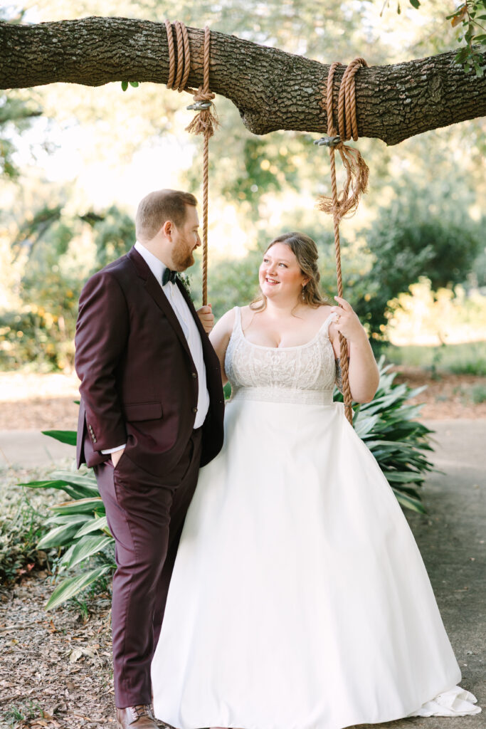 Wedding Planning in Columbia SC | Stee Garman Photography Fine-art Wedding Photographer | A couple stands by a rustic swing under a large tree. The bride smiles in a white dress while the groom in a burgundy suit gazes warmly at her.