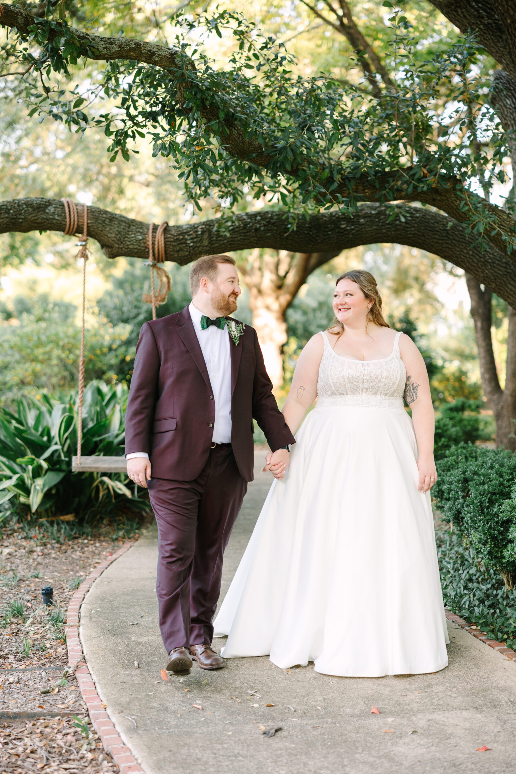 Hampton Preston Mansion Wedding | Stee Garman Photography Fine-art Wedding Photographer | A couple walks hand in hand on a garden path under trees. The groom wears a burgundy suit; the bride is in a white gown, both smiling warmly.