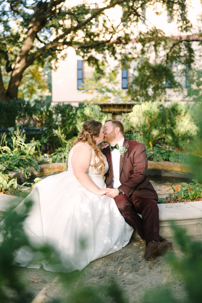 Wedding Venues in Columbia SC | Stee Garman Photography Fine-art Wedding Photographer | Bride in a white dress and groom in a maroon suit share a kiss while sitting by a garden fountain. Sunlit greenery surrounds them, creating a romantic atmosphere.