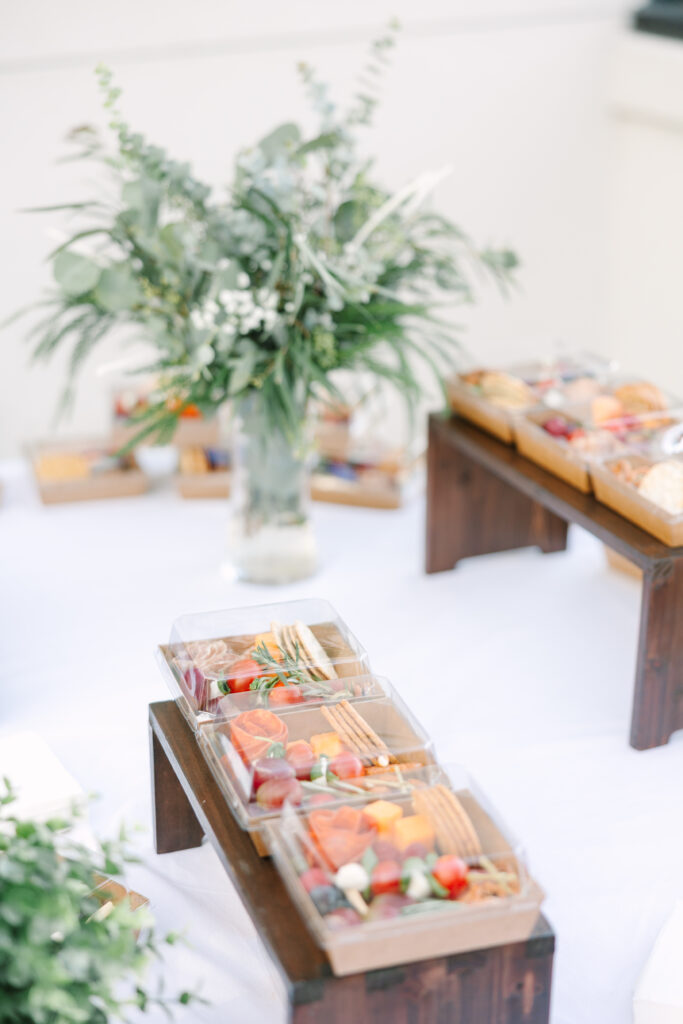 Wedding Reception Venues in Columbia SC | Stee Garman Photography Fine-art Wedding Photographer | Elegant buffet setup on a white tablecloth, featuring wooden trays with assorted snacks like cheese and fruit. A lush green floral bouquet adds a fresh touch.