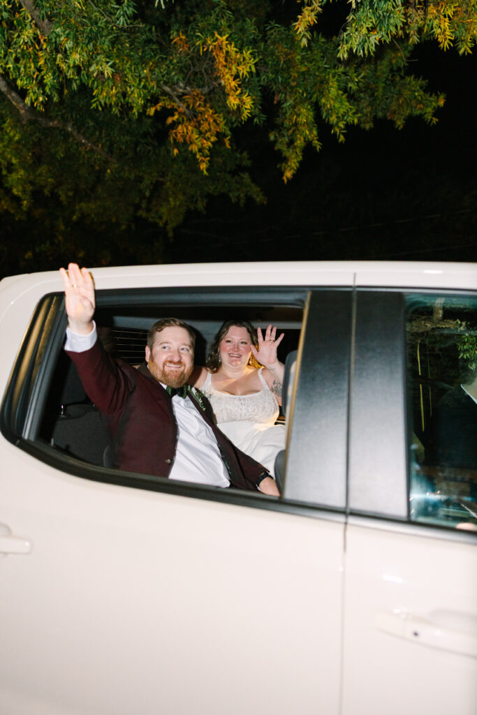 Hampton Preston Mansion Wedding | Stee Garman Photography Fine-art Wedding Photographer | Smiling couple in wedding attire waves from a car window at night, surrounded by green foliage. They exude joy and celebration.