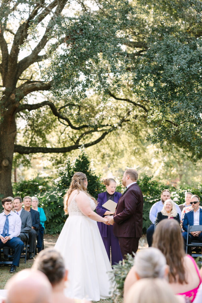 Wedding Venues in Columbia SC | Stee Garman Photography Fine-art Wedding Photographer | A couple holds hands during an outdoor wedding ceremony beneath a large tree. Guests are seated nearby, and the scene conveys joy and celebration.