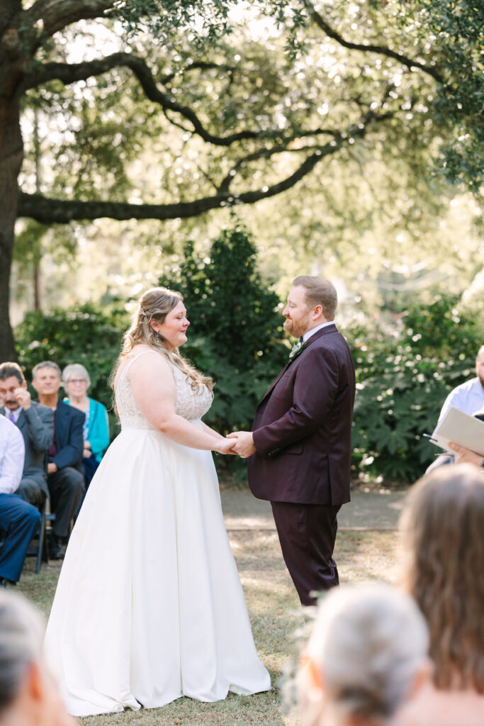 Hampton Preston Mansion Wedding | Stee Garman Photography Fine-art Wedding Photographer | Bride and groom holding hands during an outdoor wedding ceremony. They stand under a tree, surrounded by seated guests, with a serene and joyful atmosphere.