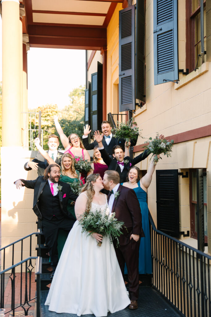 Wedding Reception Venues in Columbia SC | Stee Garman Photography Fine-art Wedding Photographer | A joyful wedding scene shows a couple kissing in the foreground, surrounded by an excited wedding party. Everyone is smiling, holding bouquets, and dressed in colorful attire, conveying a festive mood.