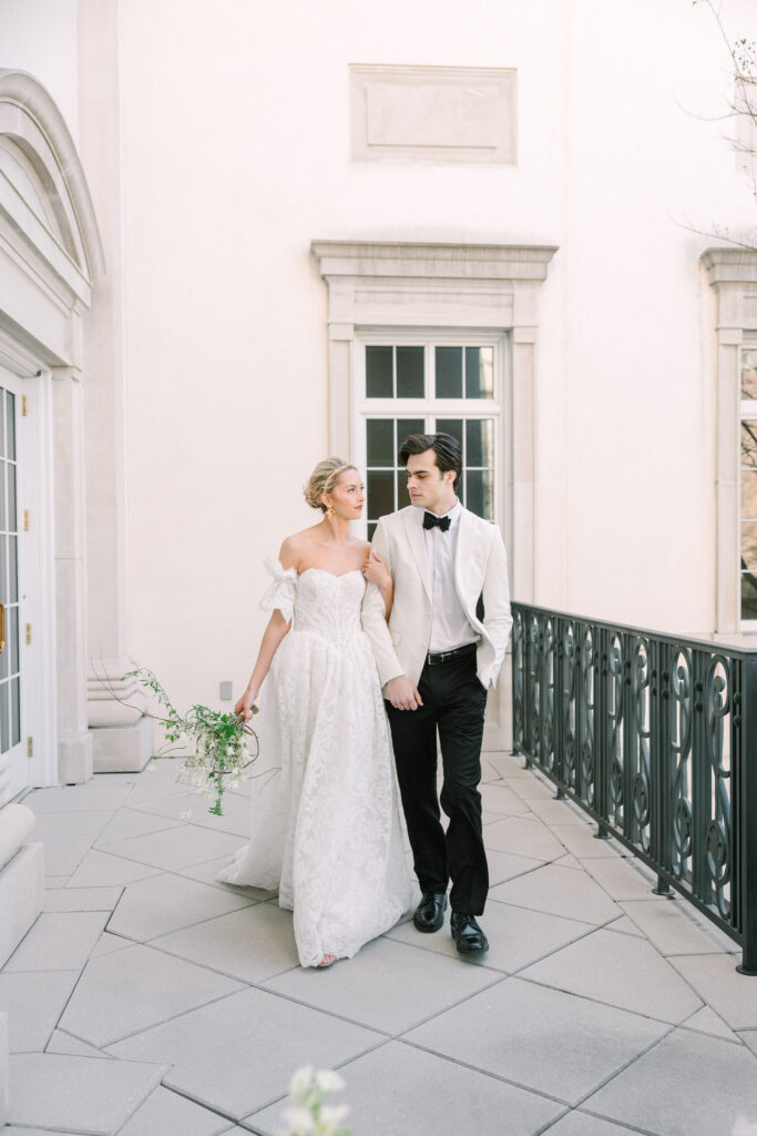 Newlyweds walk hand in hand along a grand exterior balcony with pale stone and elegant columns, a refined moment for Charleston wedding photographers.