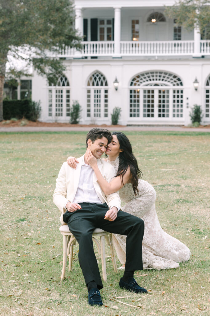 Woman leans affectionately toward her seated partner on a lawn framed by an elegant historic home, creating a refined portrait for Charleston South Carolina engagement photos.